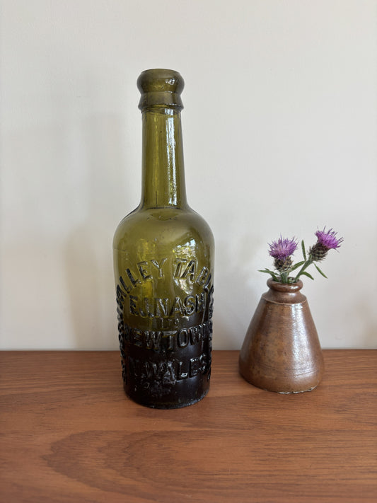 An vintage olive green glass bottle with embossed with the lettering Severn Valley Table Water, displayed on a wooden surface next to a small brown ceramic vase with purple flowers against a plain off-white wall.