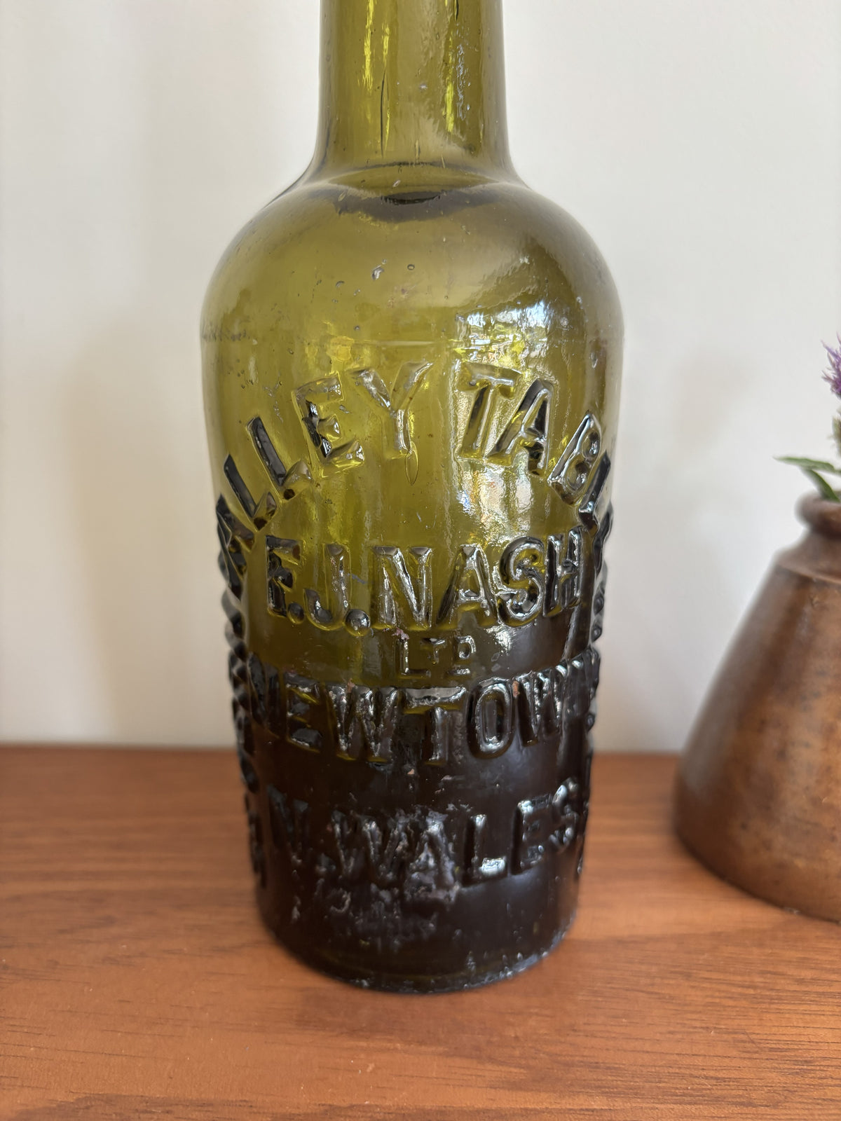 An vintage olive green glass bottle with embossed with the lettering Severn Valley Table Water, displayed on a wooden surface next to a small brown ceramic vase with purple flowers against a plain off-white wall.