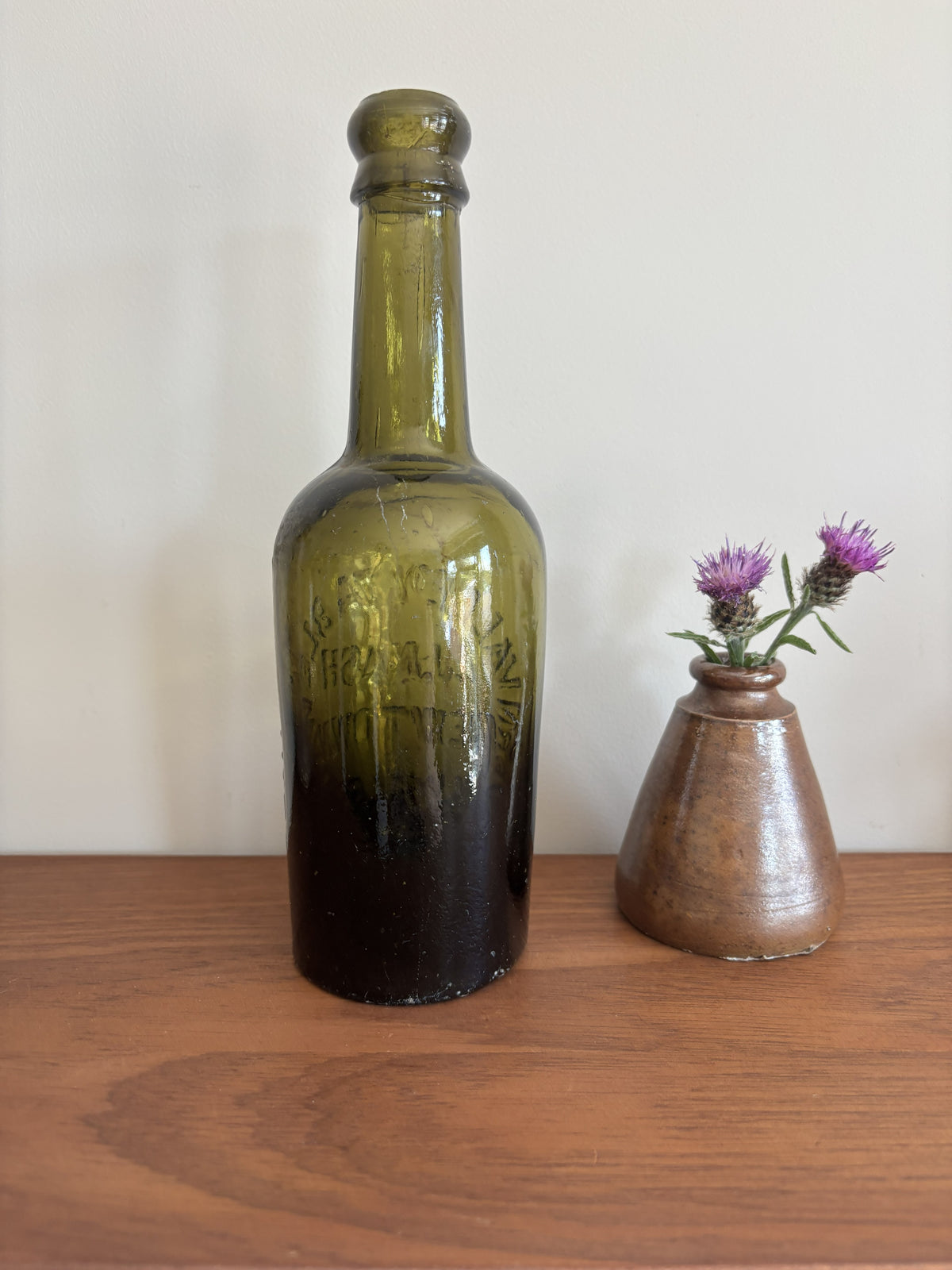 An vintage olive green glass bottle with embossed with the lettering Severn Valley Table Water, displayed on a wooden surface next to a small brown ceramic vase with purple flowers against a plain off-white wall.