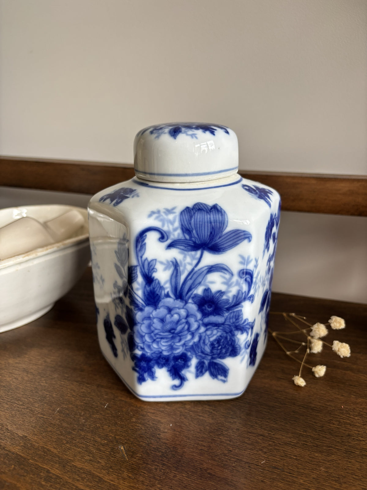 A small white ceramic ginger jar with a lid, decorated with blue floral patterns, sitting on a wooden shelf next to a white bowl and dried flowers.