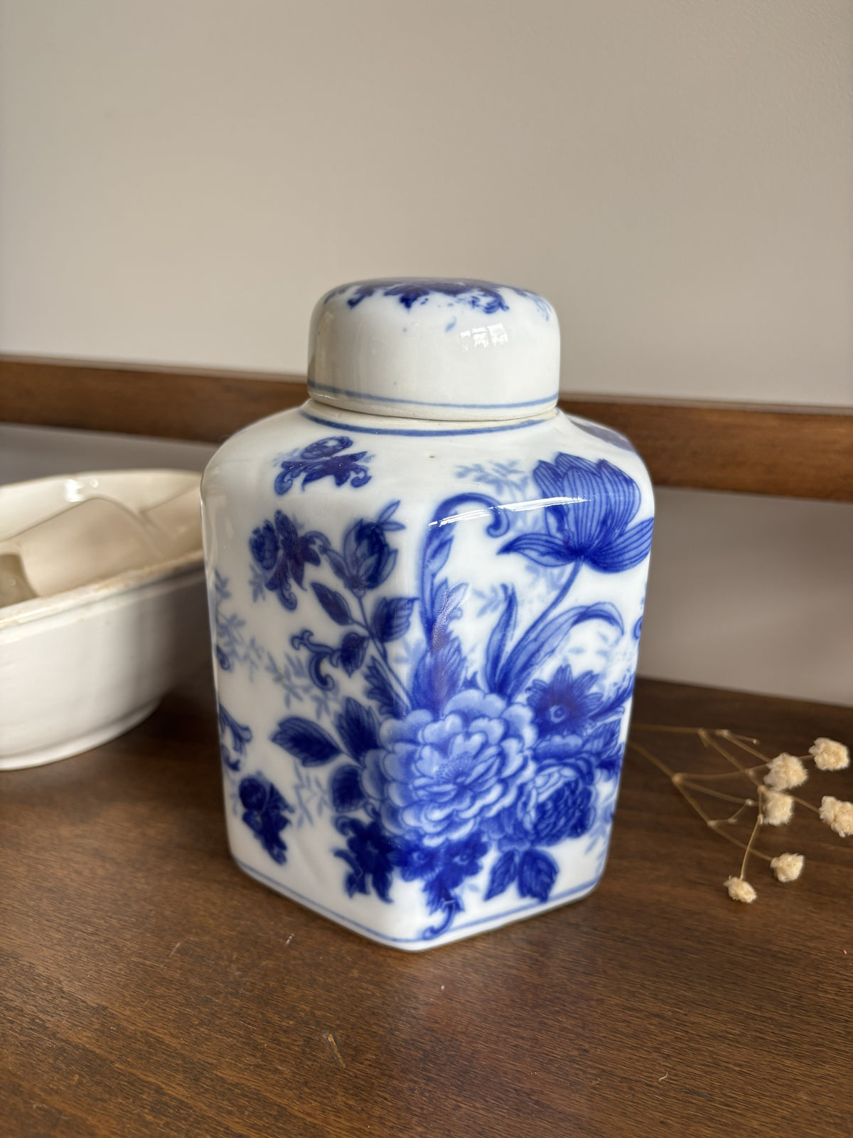 A blue and white ceramic ginger jar with a lid, decorated with a floral pattern, sitting on a wooden shelf next to a white ceramic bowl and dried flowers.