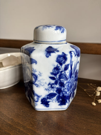 A hexagonal white ceramic ginger jar with a matching lid, decorated with a classic blue floral pattern, sitting on a wooden surface.