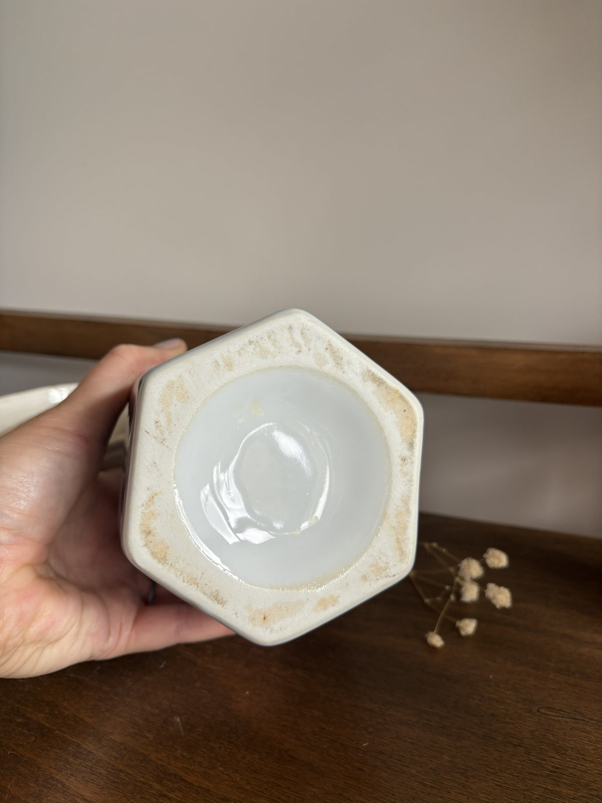 A hand holding the bottom of a hexagonal ceramic ginger jar, showing a glossy off-white glazed interior with a rustic, lightly stained beige rim, set against a dark wooden shelf background.