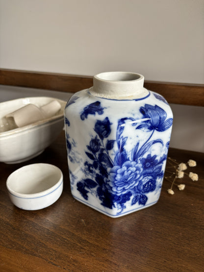 A hexagonal ceramic ginger jar with a matching small cup, decorated in a classic blue and white floral pattern, displayed on a wooden table.