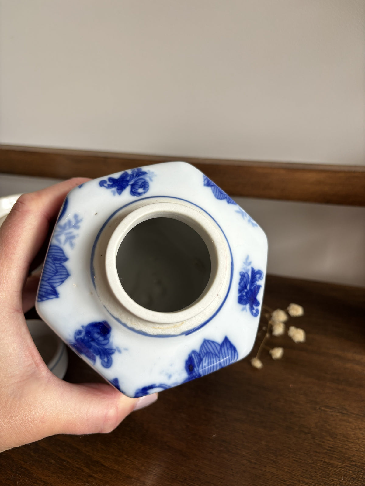 A hand holding a hexagonal white ceramic ginger jar with blue floral patterns, showing the interior opening, set on a wooden surface.