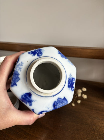 A hand holding a hexagonal white ceramic ginger jar with blue floral patterns, showing the interior opening, set on a wooden surface.
