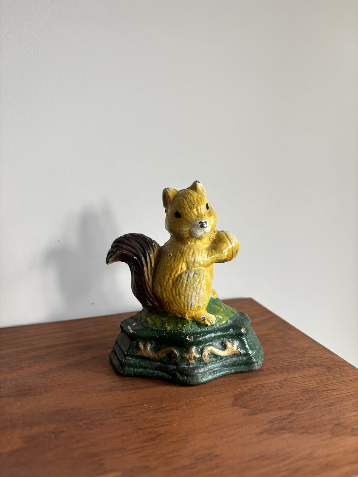 A small cast iron squirrel cast iron doorstop in yellow and dark brown, standing on a green and gold base, placed on a wooden surface against a plain light gray wall.