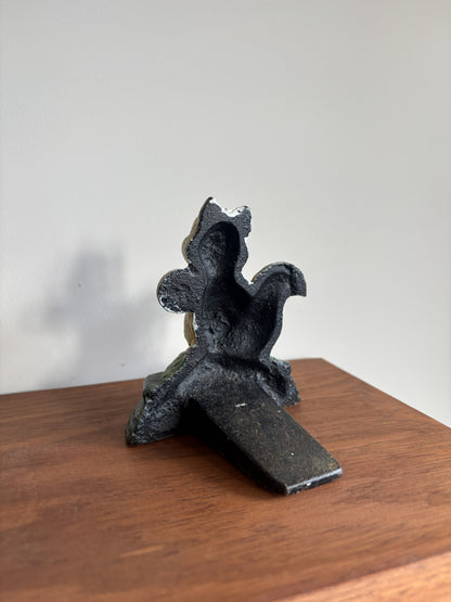 The back of a small cast iron door stop with a textured, weathered finish, standing on a wooden surface against a plain light gray wall.