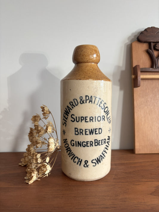A vintage ceramic bottle with a brown and cream glaze, featuring printed text that reads 'Steward & Patteson Superior Brewed Ginger Beer Norwich & Swaffham'. The bottle is displayed on a wooden surface next to dried flowers against a plain wall.