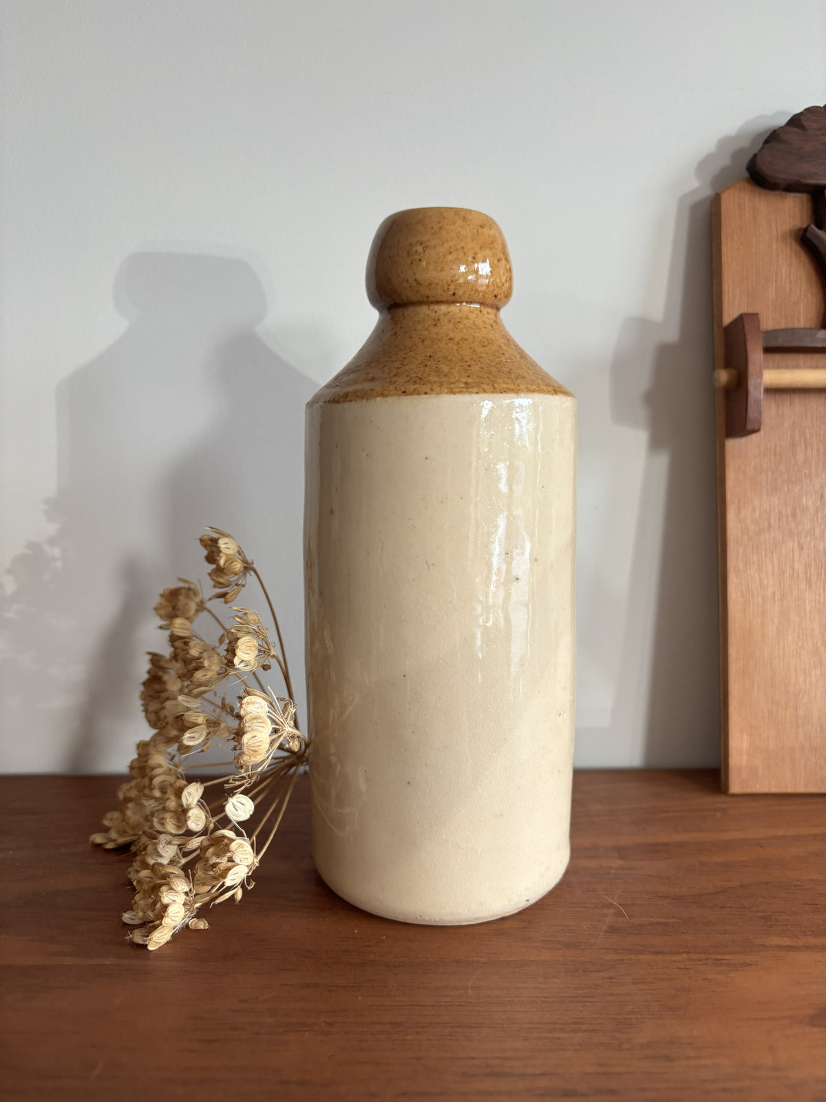 A ceramic bottle with a textured brown glaze on the neck and a matte cream-colored body, displayed on a wooden surface next to dried flowers against a plain wall.
