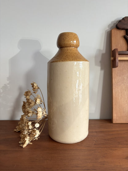 A ceramic bottle with a textured brown glaze on the neck and a matte cream-colored body, displayed on a wooden surface next to dried flowers against a plain wall.