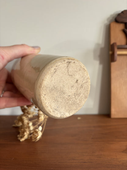 A hand holding a cylindrical ceramic bottle upside down to show the textured, unglazed bottom, set on a wooden table with a neutral background.