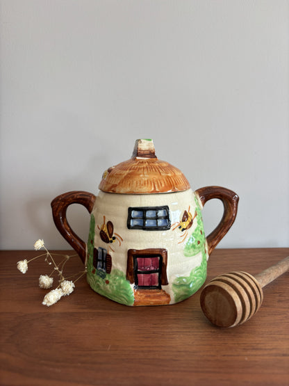 A Suvesco ceramic honey pot with a house-shaped body, brown handles, and a brown lid, decorated with painted bees and a pink door.