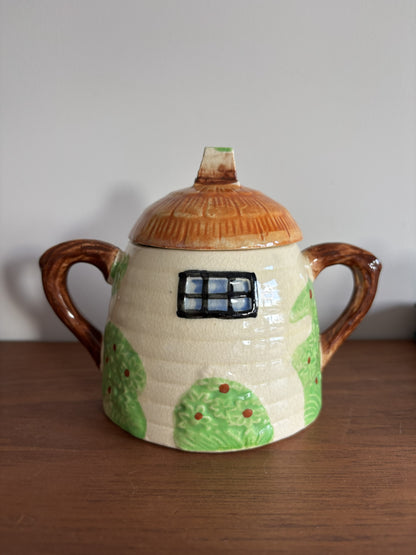 A Suvesco ceramic honey pot with a house-shaped body, brown handles, and a brown lid, decorated with a painted window.  