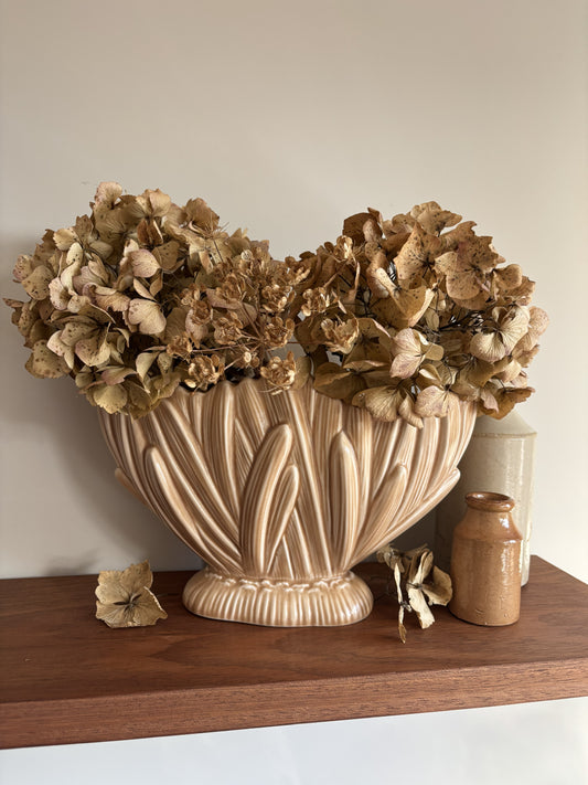 A fawn coloured vintage SylvaC mantel vase filled with dried beige hydrangea flowers, displayed on a dark wood shelf next to a small brown bottle and a cream vase against a neutral wall.