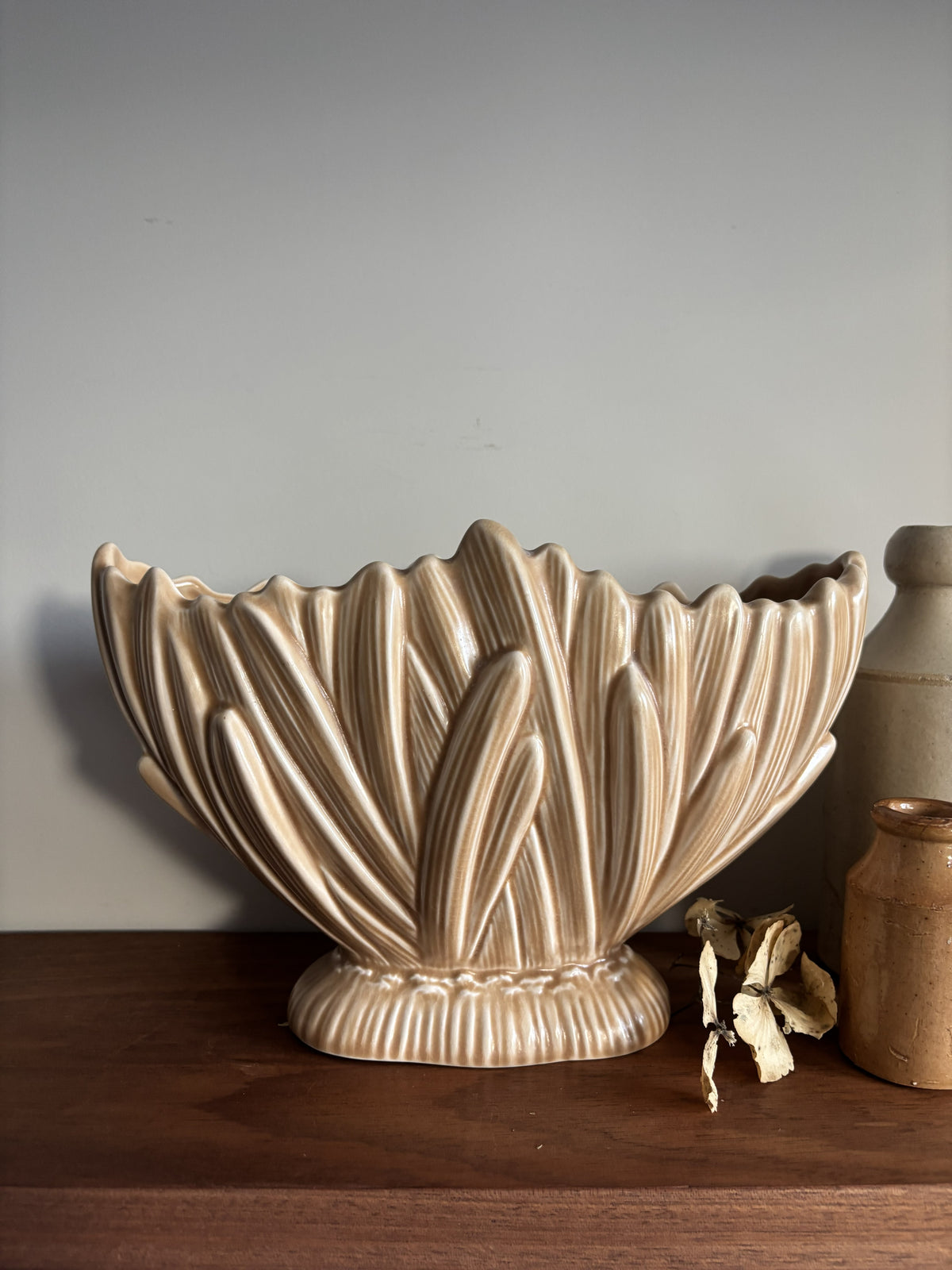 A fawn coloured vintage SylvaC mantel vase  displayed on a dark wood shelf next to a small brown bottle and a cream vase against a neutral wall.