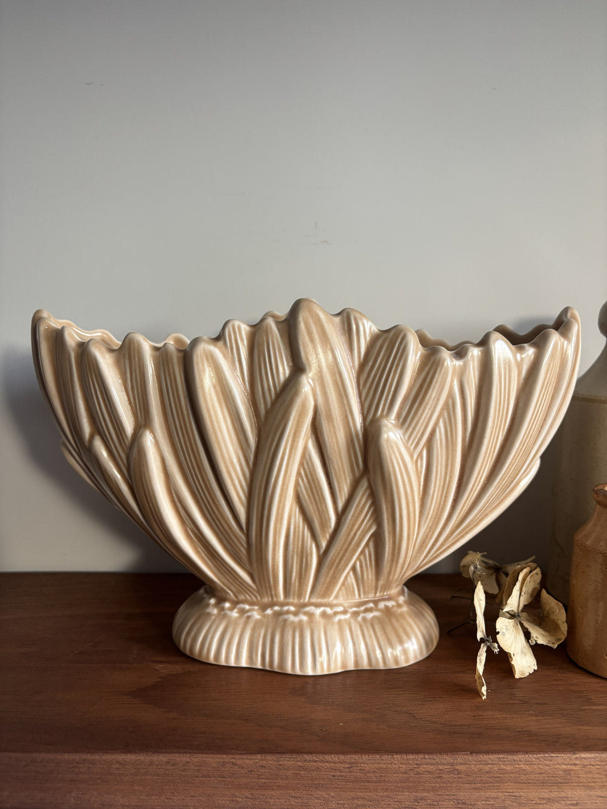 A fawn coloured vintage SylvaC mantel vase  displayed on a dark wood shelf next to a small brown bottle and a cream vase against a neutral wall.