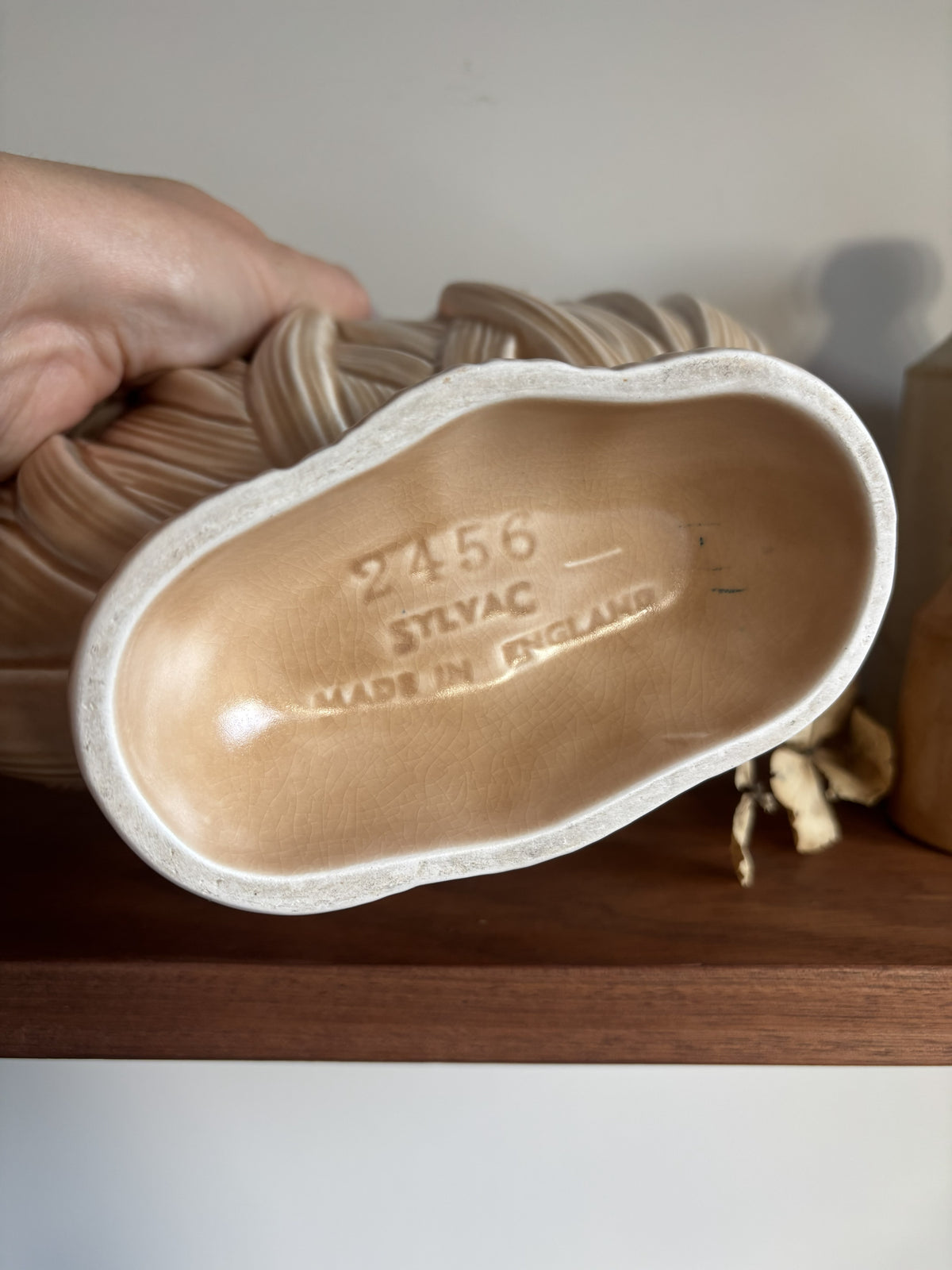 A hand holding the bottom of a fawn ceramic coloured vase showing the embossed text '2456 SYLYAC MADE IN ENGLAND' on a wooden shelf