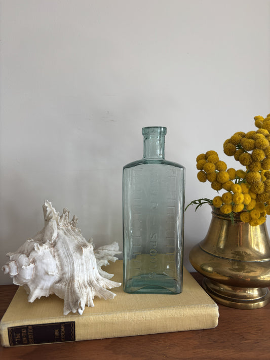 A clear glass bottle with embossed measurement markings and the words Table Spoons standing on a vintage cream-colored book next to a white seashell and yellow flowers in a brass pot against a plain wall.
