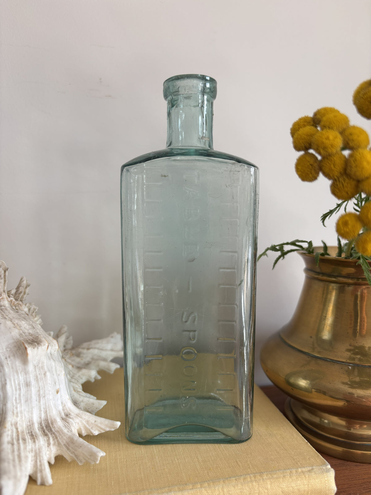 A clear glass bottle with embossed measurement markings and the words Table Spoons standing on a vintage cream-colored book next to a white seashell and yellow flowers in a brass pot against a plain wall.