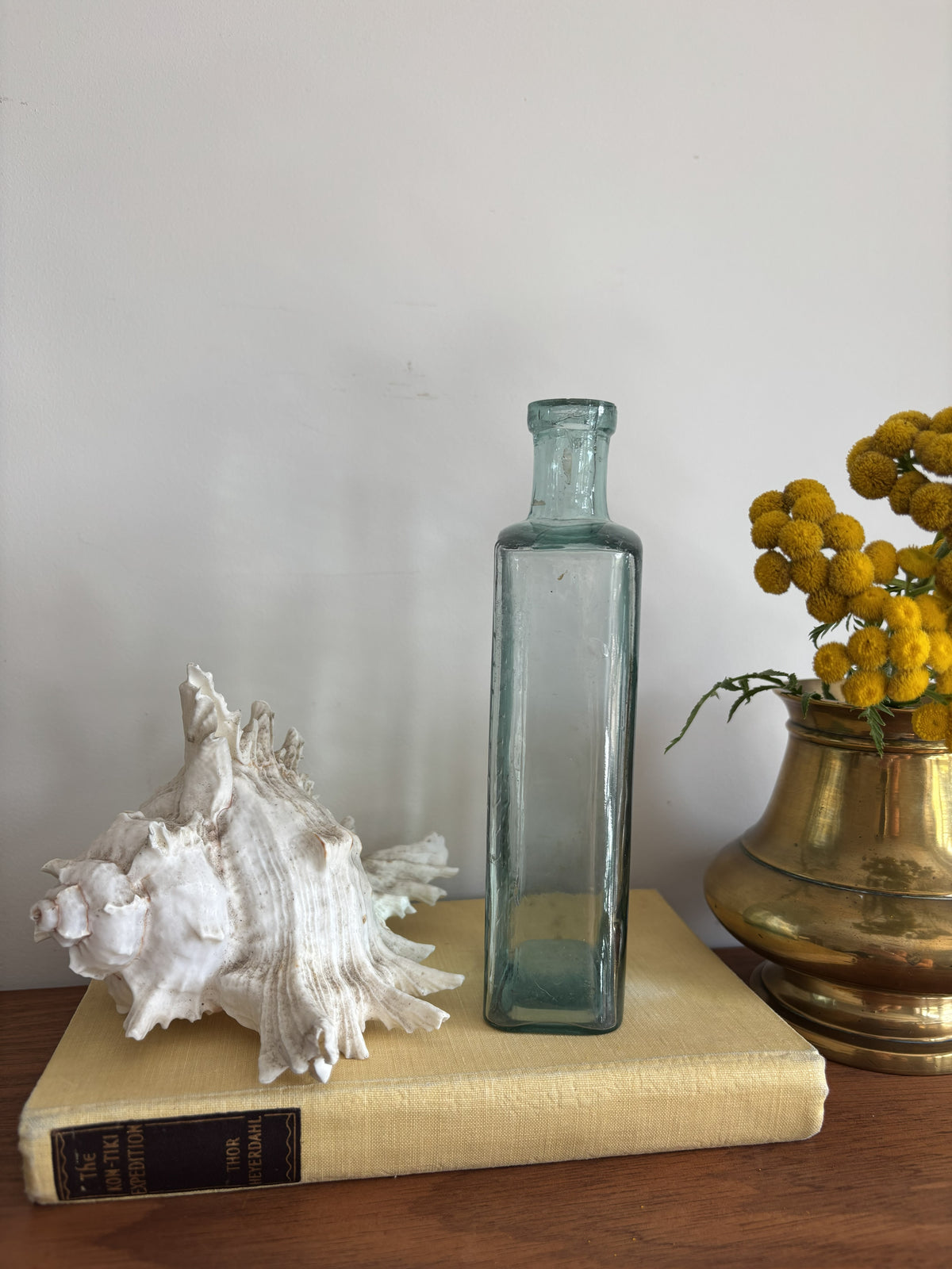 A clear glass bottle with embossed measurement markings and the words Table Spoons standing on a vintage cream-colored book next to a white seashell and yellow flowers in a brass pot against a plain wall.