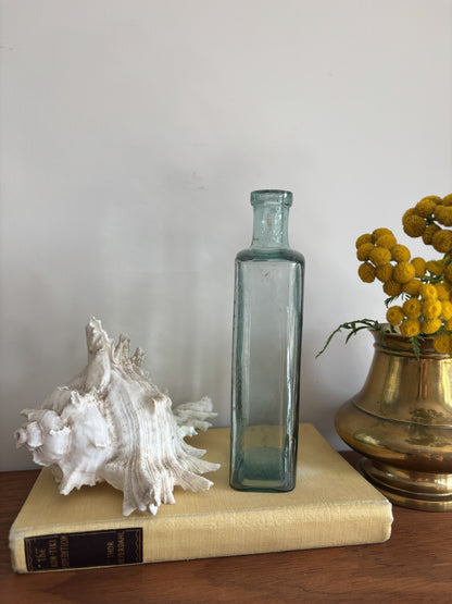 A clear glass bottle with embossed measurement markings and the words Table Spoons standing on a vintage cream-colored book next to a white seashell and yellow flowers in a brass pot against a plain wall.