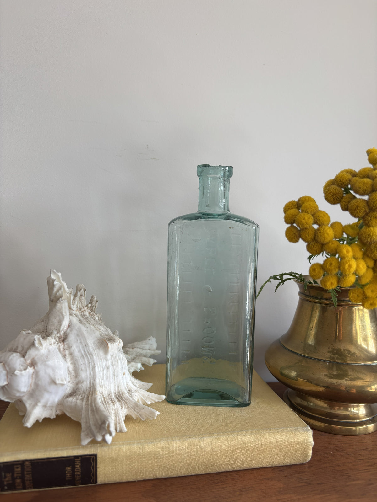 A clear glass bottle with embossed measurement markings and the words Table Spoons standing on a vintage cream-colored book next to a white seashell and yellow flowers in a brass pot against a plain wall.
