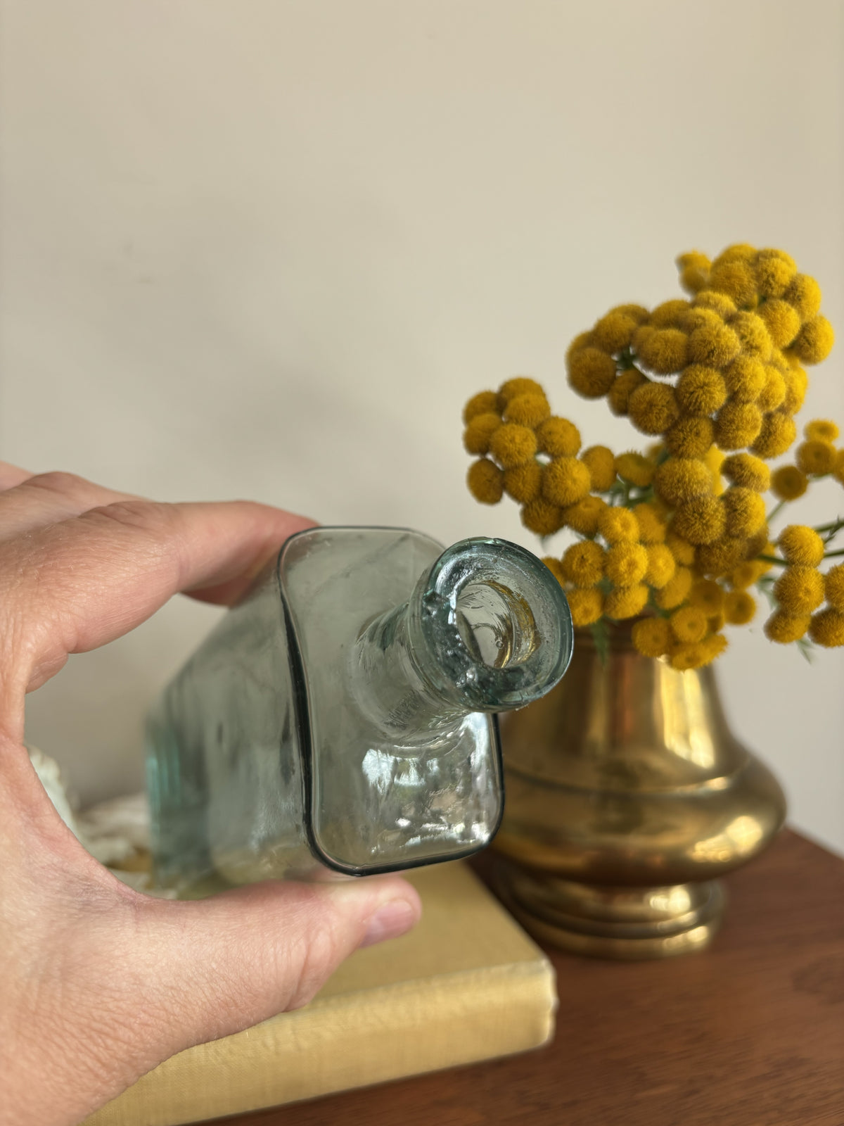 A clear glass bottle with embossed measurement markings and the words Table Spoons standing on a vintage cream-colored book next to a white seashell and yellow flowers in a brass pot against a plain wall.
