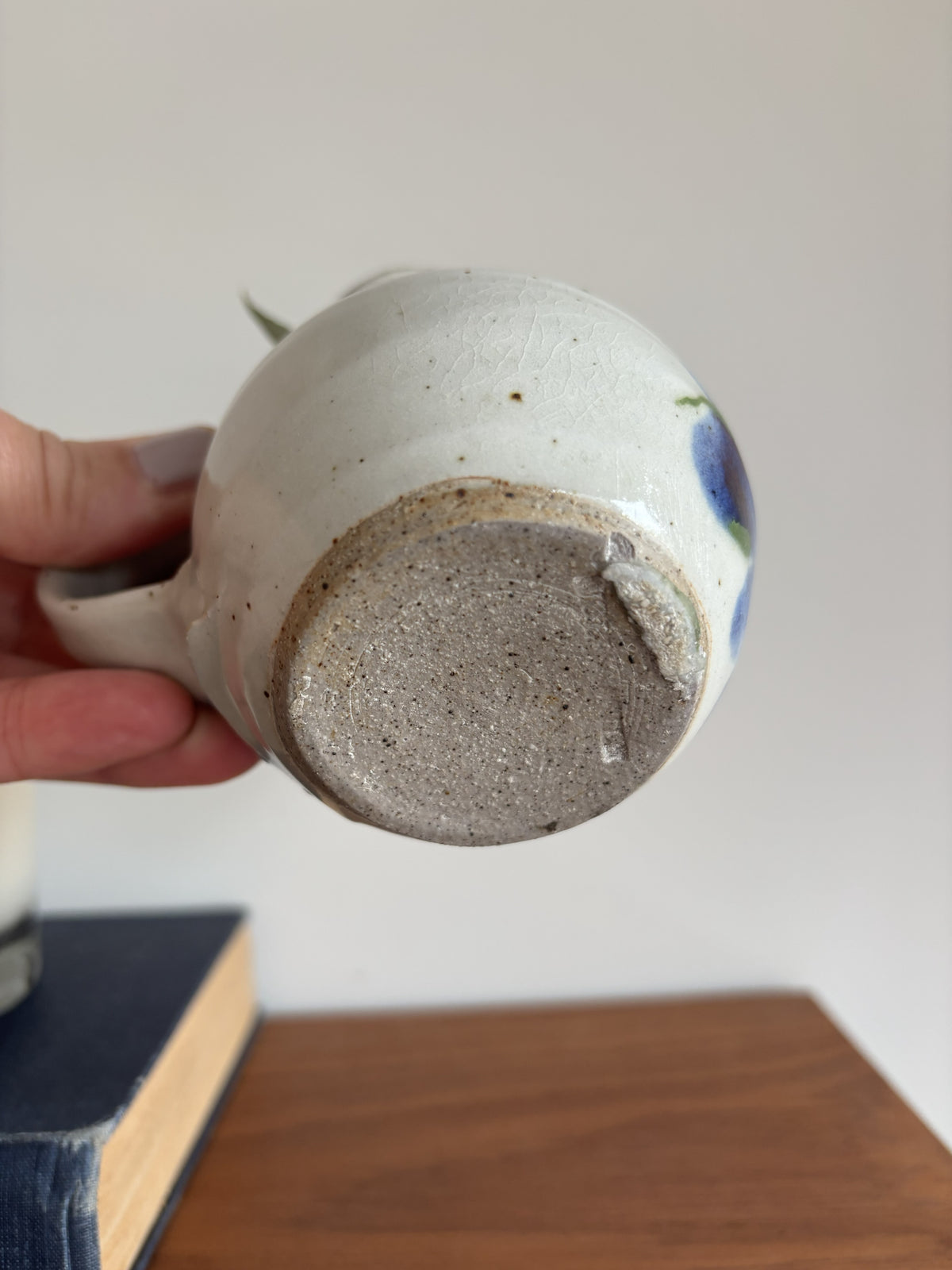 A hand holds a handmade ceramic mug upside down to show the textured, speckled clay base. The mug has a glossy off-white glaze with a blue and green floral design on the side and is photographed against a neutral indoor background with a wooden table surface.