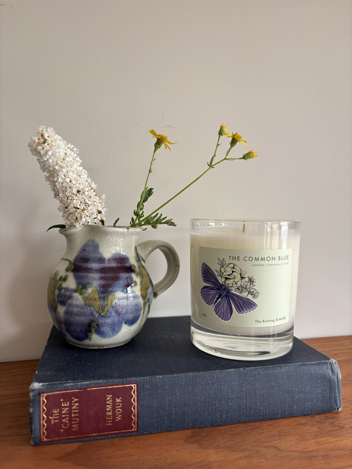 A glass jar candle labeled 'THE COMMON BLUE' sits on a navy blue book next to a ceramic pitcher filled with white and yellow flowers, set against a plain light gray wall.