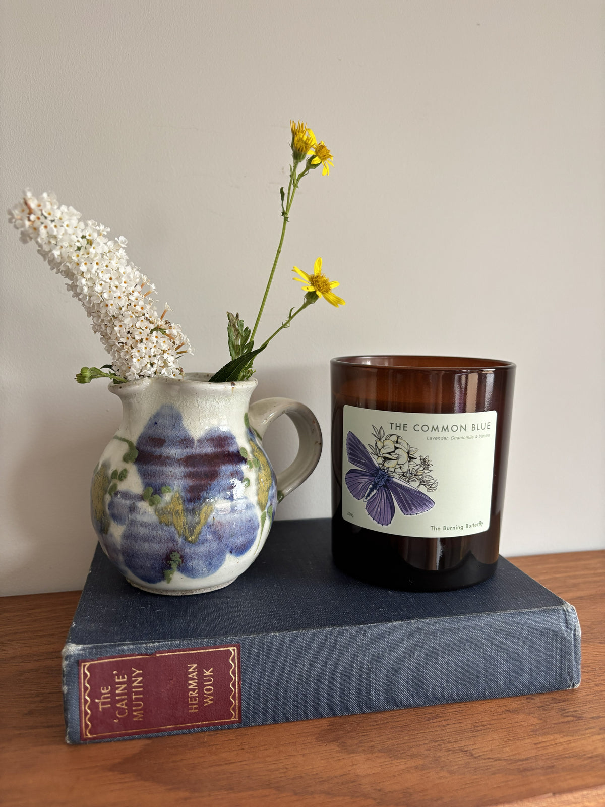 An amber glass candle jar with a white label reading 'THE COMMON BLUE' and a purple butterfly illustration, styled on top of a blue book next to a ceramic pitcher with white and yellow flowers against a plain wall.