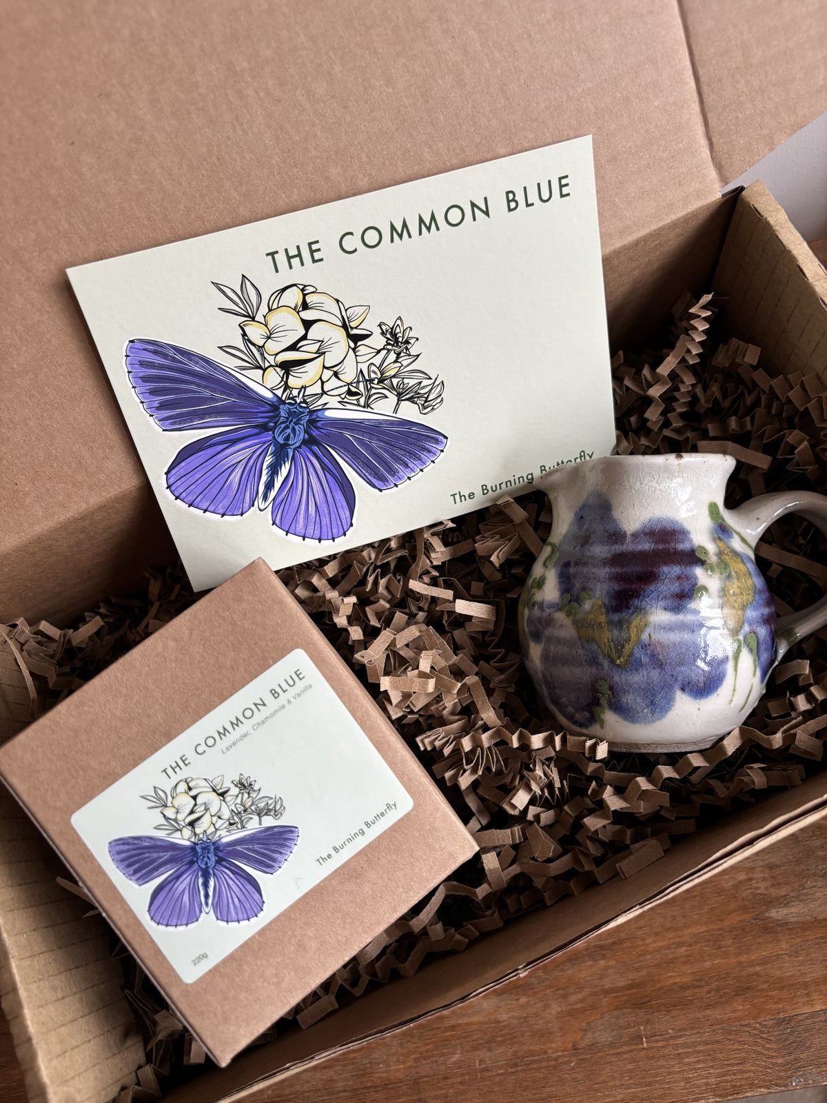 A gift box from The Burning Butterfly containing The Common Blue candle in a kraft gift box, a small blue and white bud vase and The Common Blue butterfly A6 postcard. 