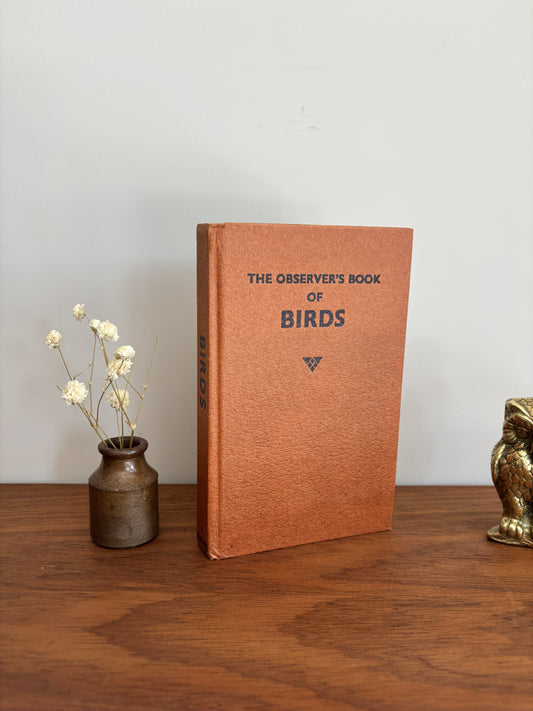 The Observer's Book of Birds book with a textured orange-brown cover standing on a wooden table, flanked by a small vase of dried flowers on the left and a brass owl figurine on the right, against a plain light gray wall.