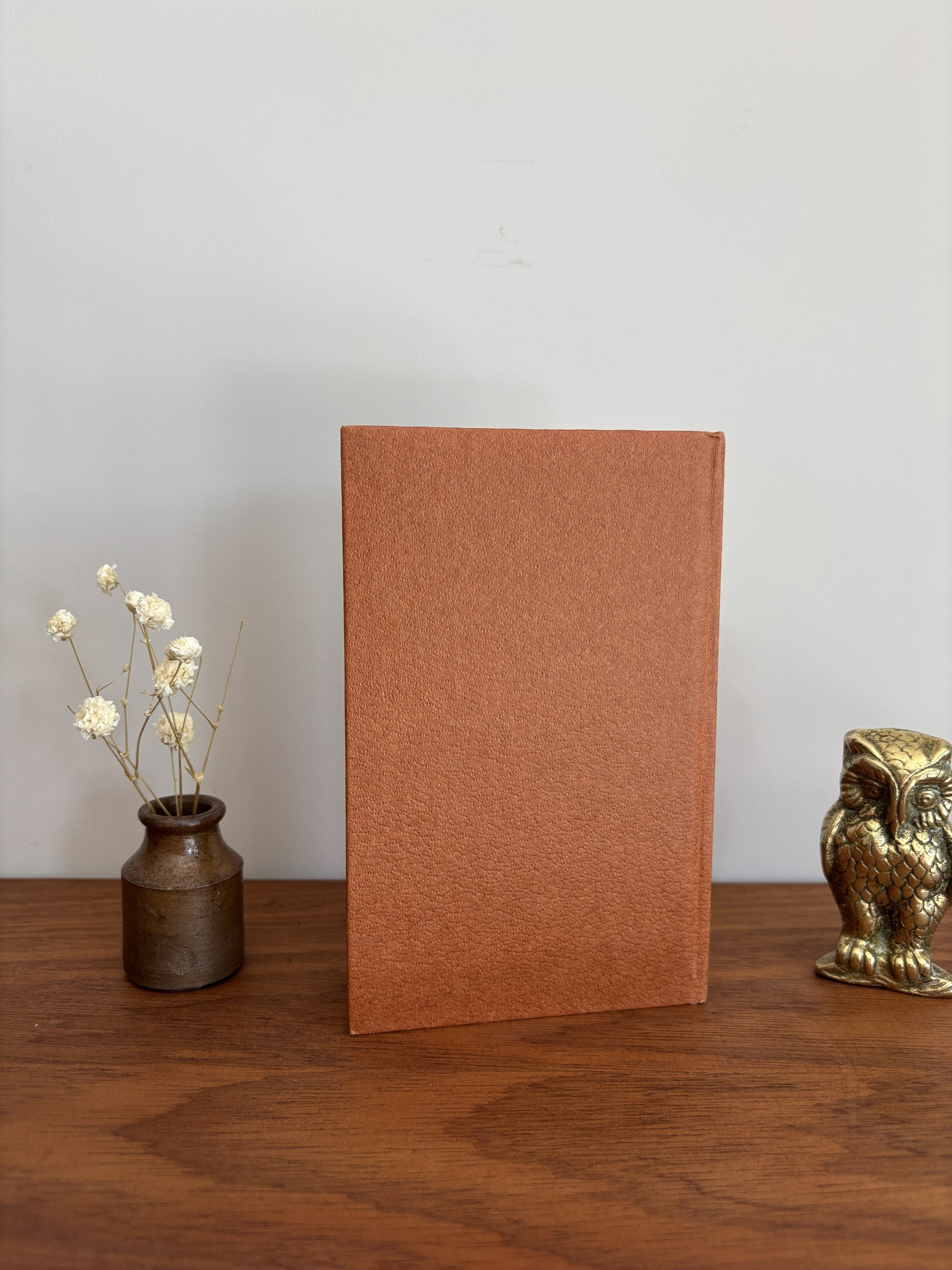A textured, rectangular terracotta-brown book or notebook standing upright on a wooden table, flanked by a small vase with dried white flowers on the left and a small metallic owl figurine on the right, against a plain light gray wall.