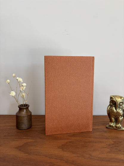 A textured, rectangular terracotta-brown book or notebook standing upright on a wooden table, flanked by a small vase with dried white flowers on the left and a small metallic owl figurine on the right, against a plain light gray wall.