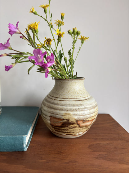 A handmade ceramic vase with a textured beige and brown glaze, filled with yellow daisies and purple flowers, sitting on a wooden table next to a teal book against a plain white wall.