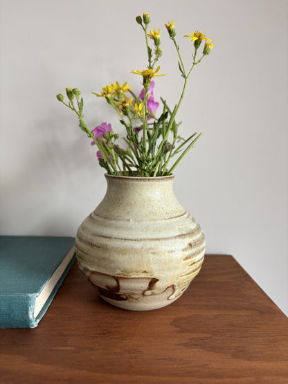 A handmade ceramic vase with a textured, speckled beige and brown glaze, filled with wildflowers, sitting on a wooden table next to a teal book against a neutral gray wall.
