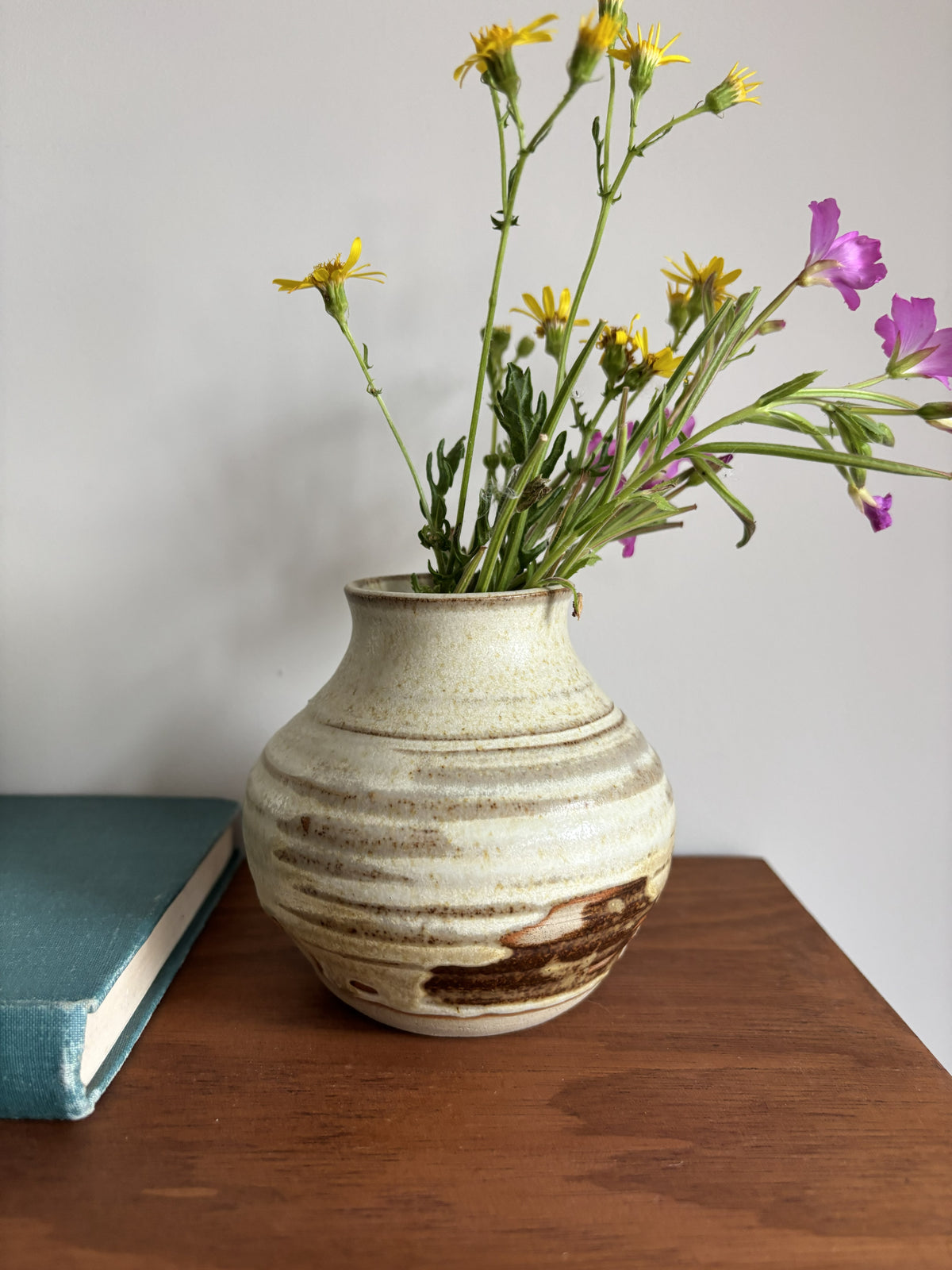 A handmade ceramic vase with a textured, ribbed surface in cream and brown tones, filled with yellow and purple wildflowers, sitting on a wooden table next to a teal book against a neutral gray wall.
