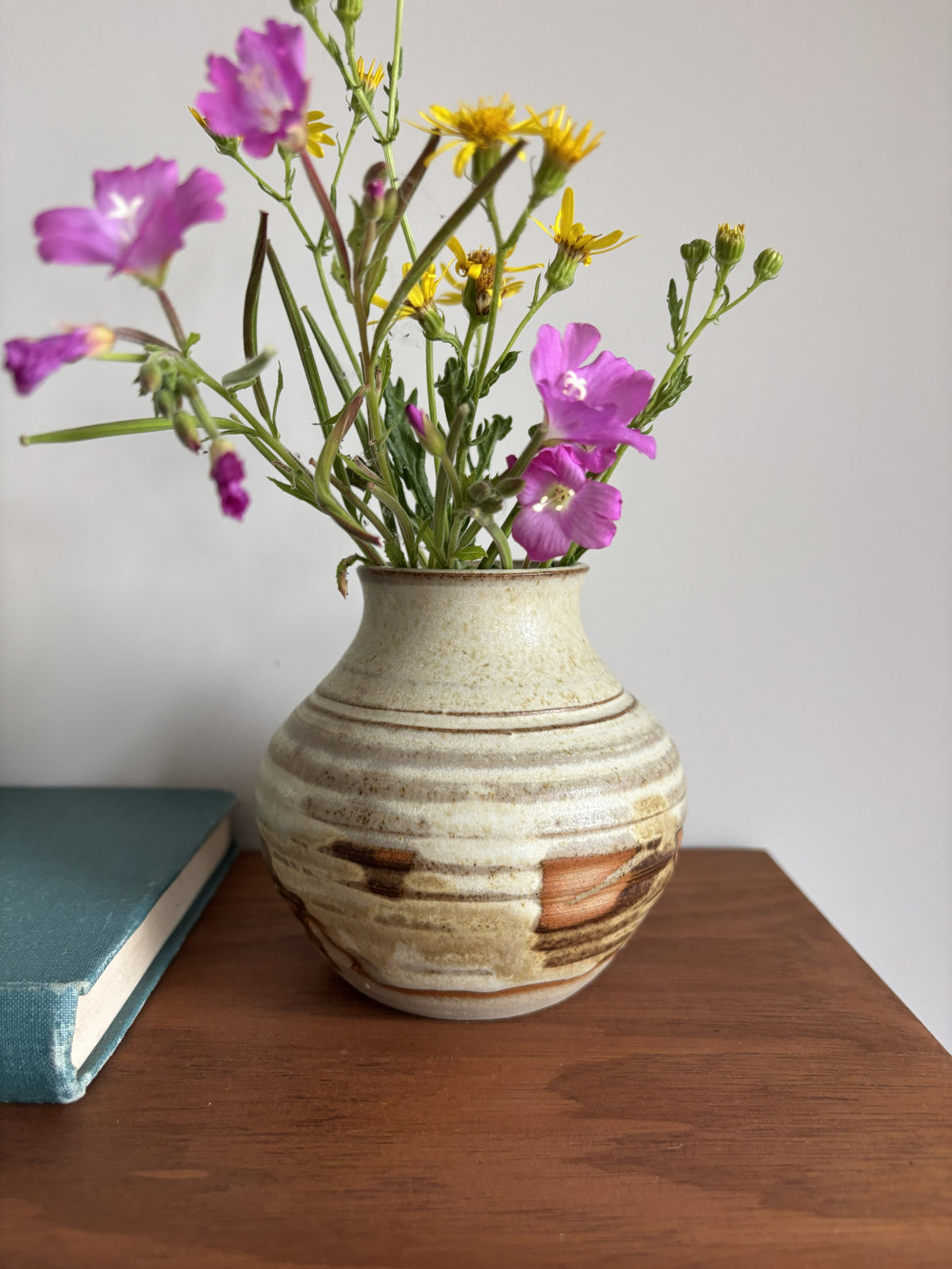 A handmade ceramic vase with a textured, speckled beige and brown glaze, filled with purple and yellow wildflowers, sitting on a wooden table next to a teal book against a plain light gray wall.