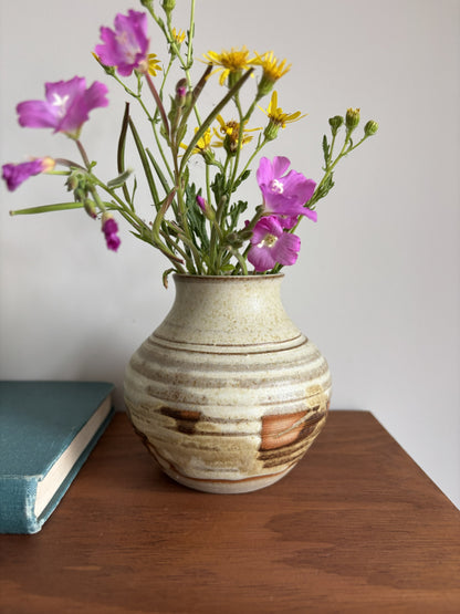 A handmade ceramic vase with a textured, speckled beige and brown glaze, filled with purple and yellow wildflowers, sitting on a wooden table next to a teal book against a plain light gray wall.