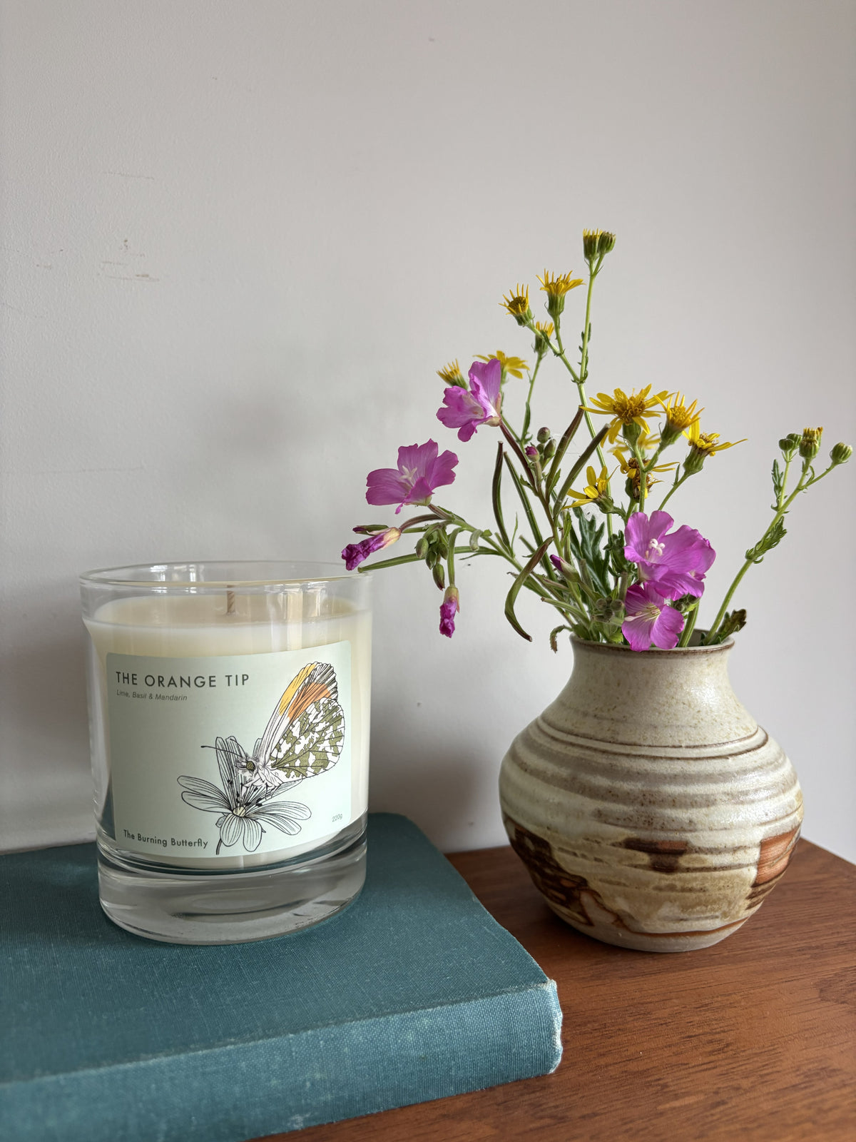 A scented candle in a clear glass jar sits on a teal book next to a ceramic vase filled with yellow and purple wildflowers, set against a plain off-white wall.