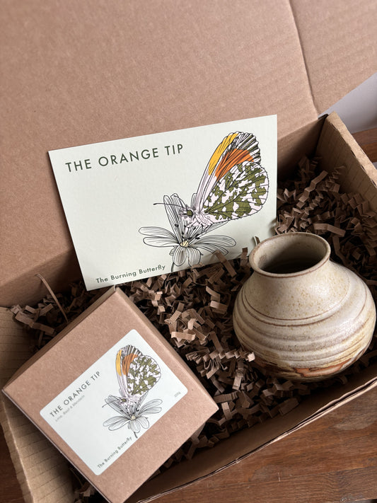 A gift box from The Burning Butterfly containing The Orange Tip candle in a kraft gift box, a small cream and orange bud vase and The Orange Tip butterfly A6 postcard. 