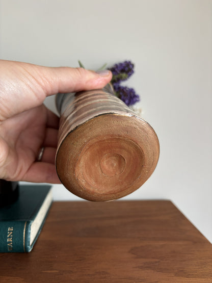 A hand holds a handmade ceramic vase with a rustic wood-grain base and a narrow metallic silver neck, containing dried purple flowers, set on a wooden table.