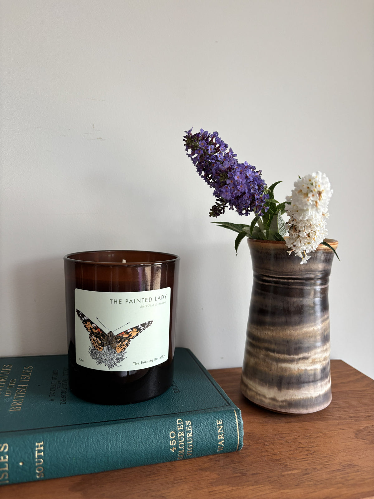 An amber glass jar candle labeled 'THE PAINTED LADY sits on a teal book next to a tall striped ceramic vase holding purple and white flowers, against a plain light wall.