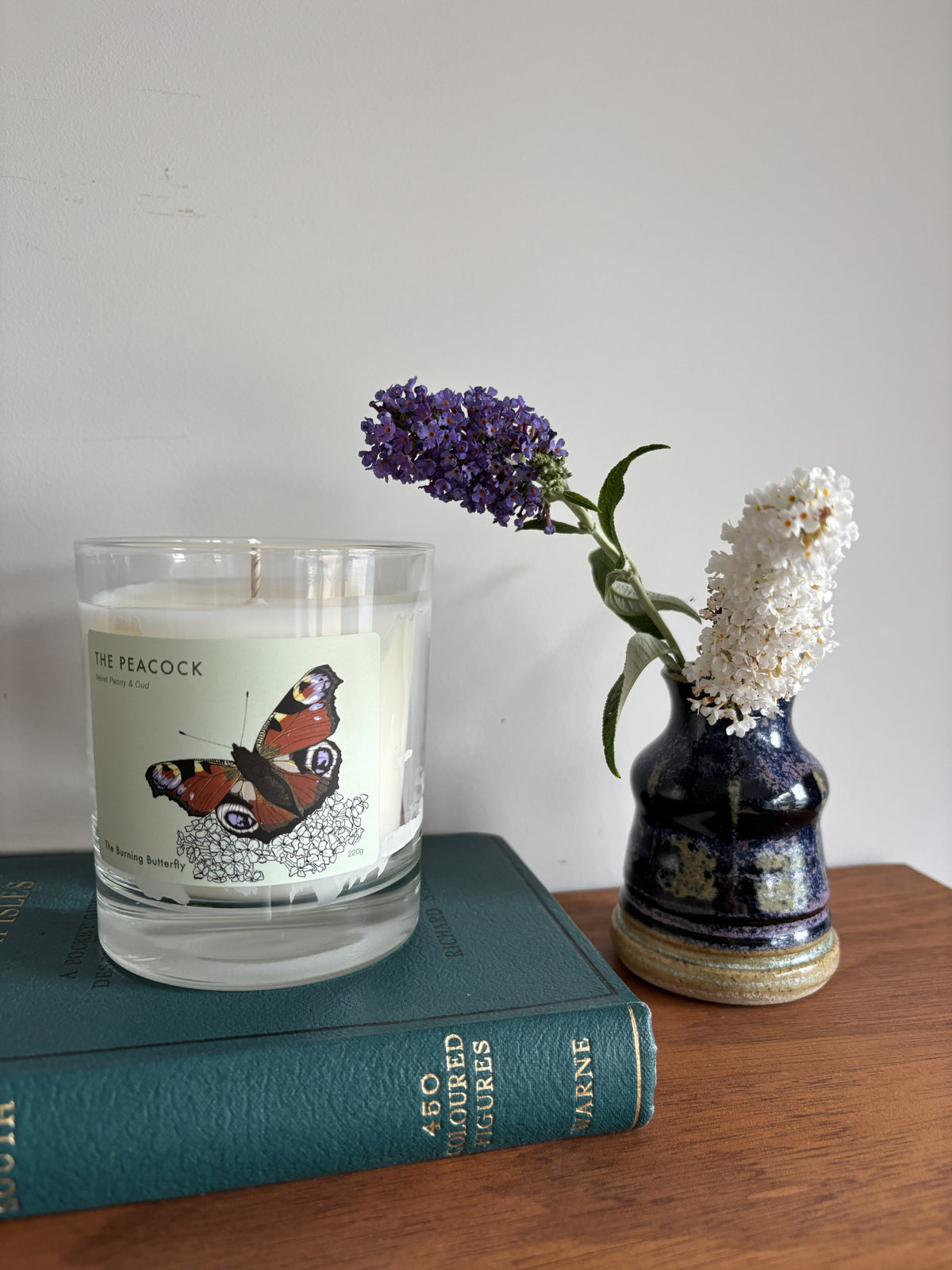 A glass jar candle with a white wax body and a butterfly label reading 'THE PEACOCK' sits on a teal book next to a small dark blue ceramic vase holding purple and white flowers, set on a wooden surface against a plain wall.