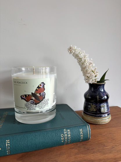 A clear glass jar candle with white wax and a butterfly label reading 'THE PEACOCK' sits on top of a teal book. To the right, a small dark blue ceramic vase holds white flowers on a wooden surface against a plain light wall.