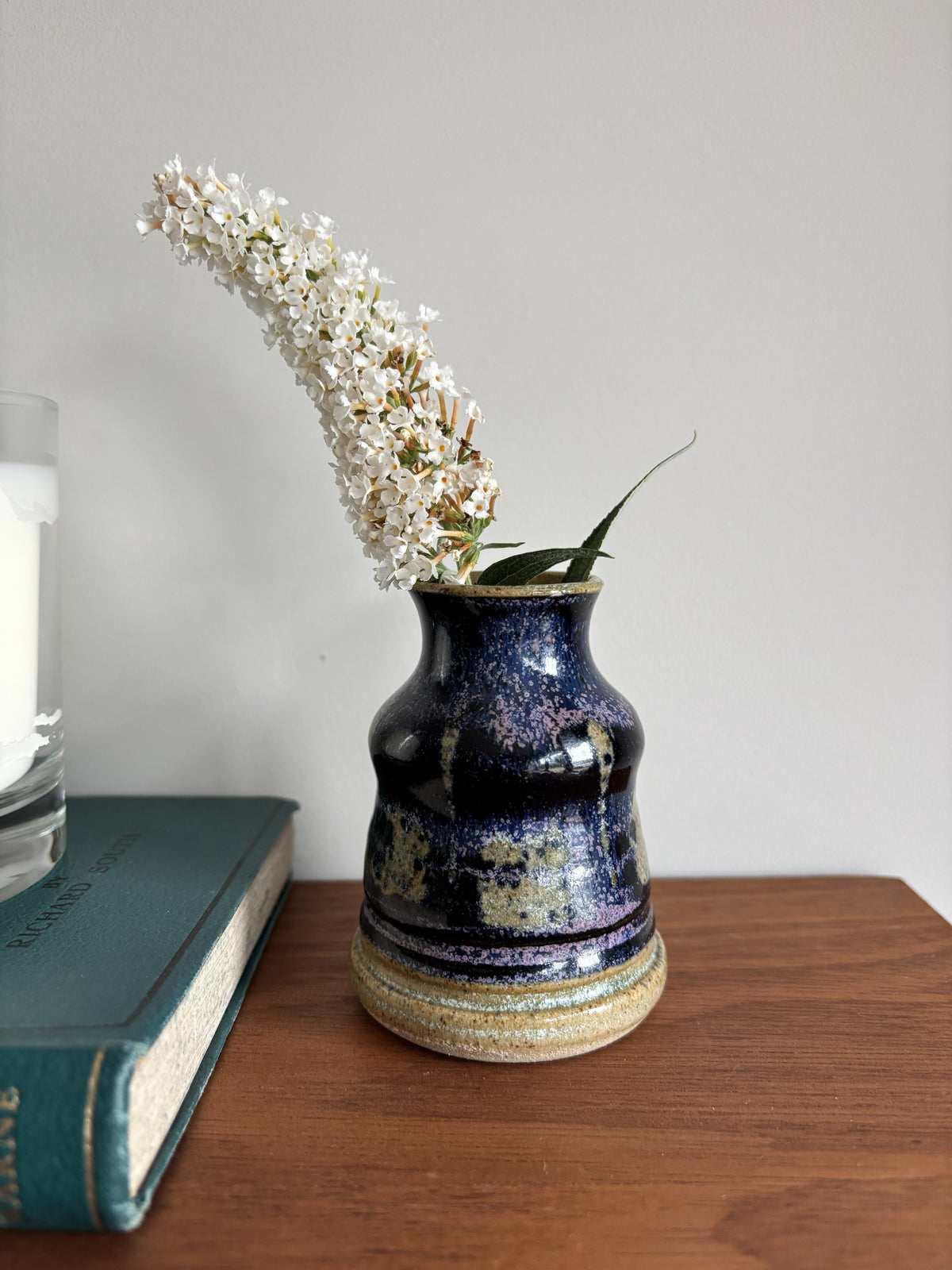 A small dark purple ceramic bud vase containing white flowers sitting on a brown shelf