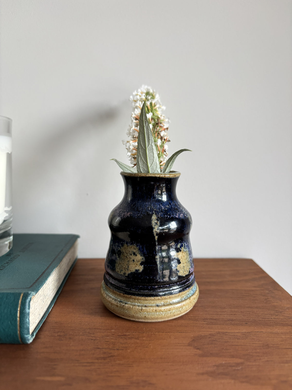 A small handmade ceramic vase with a dark blue and gold speckled glaze, holding a small white flower, sitting on a wooden table next to a book.