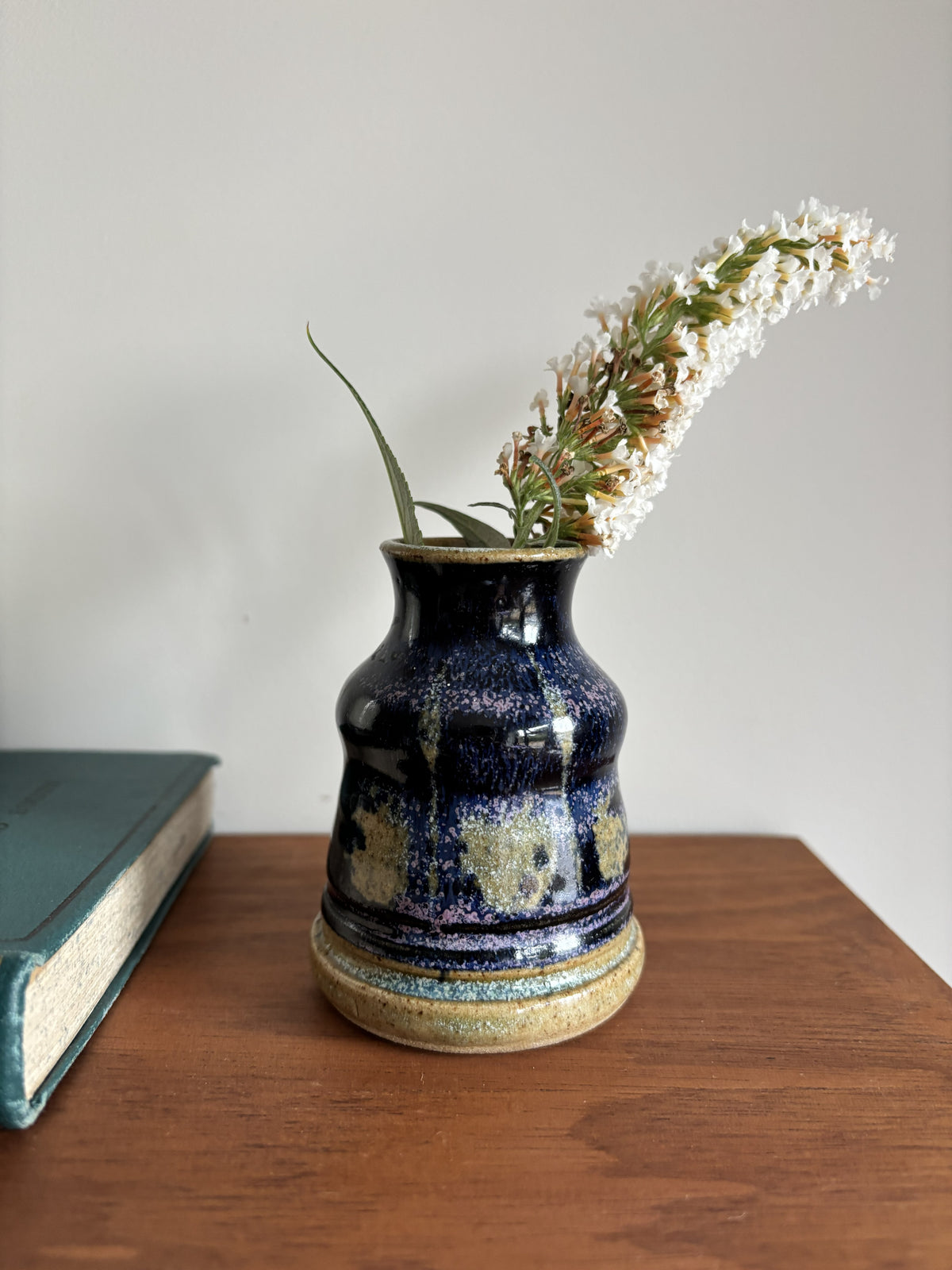 A small handmade ceramic vase with a dark blue and gold speckled glaze, holding white flowers, set on a wooden table next to a book against a plain wall.