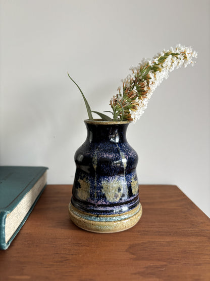 A small handmade ceramic vase with a dark blue and gold speckled glaze, holding white flowers, set on a wooden table next to a book against a plain wall.